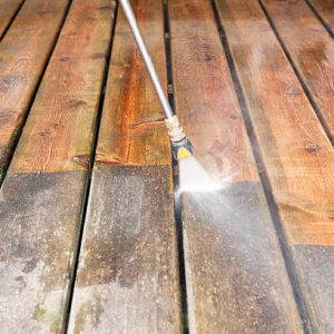 A pressure washer sprayer is cleaning a weathered treated wood deck. The background wood has been cleaned, the lower portion is dirty and weathered, showing the contrast. Focus is on the wood, the nozzle is slightly soft.