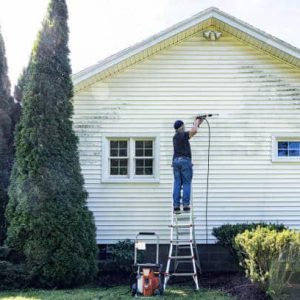 Real life, real person DIY senior man is balanced near the top of a ladder while cleaning the vinyl clapboard siding on his house with high pressure cleaning power wash equipment. Very satisfying to see such clear progress as he methodically moves the spray nozzle back and forth across the grungy grimy surface!